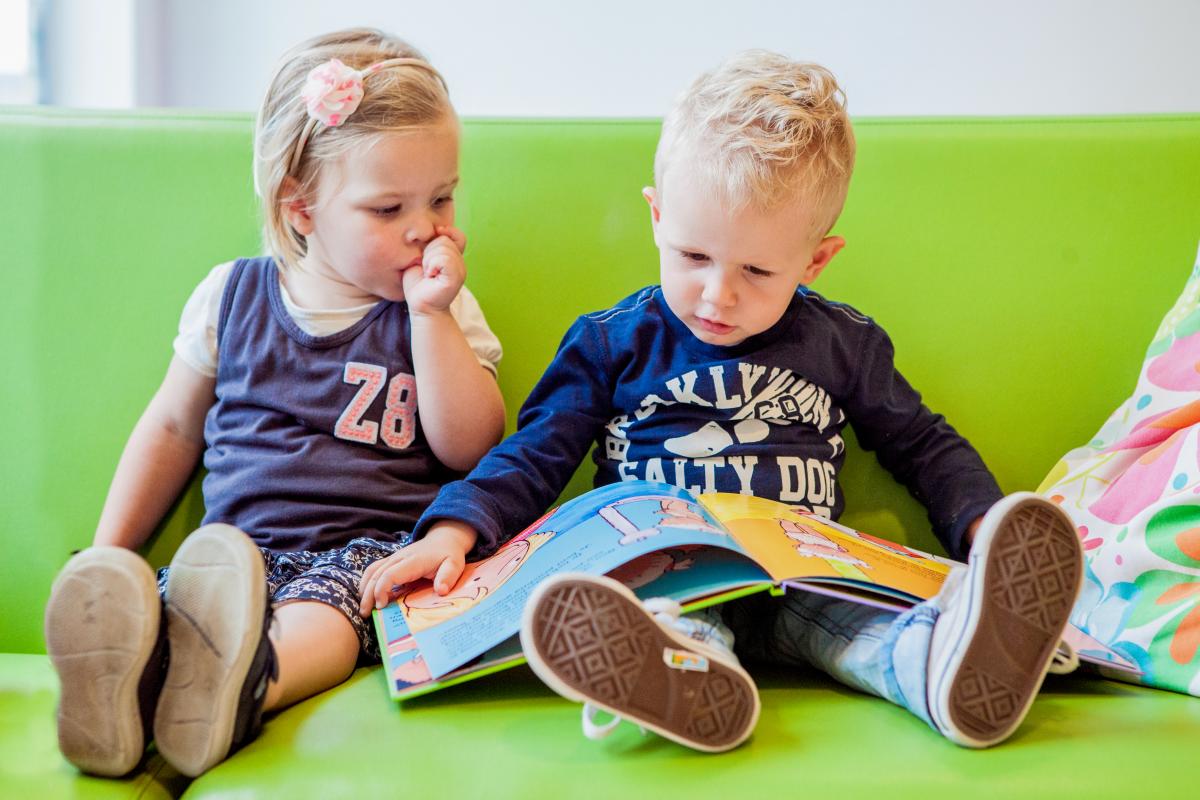 Een meisje en jongen lezen samen een boek op de bank van het kinderdagverblijf